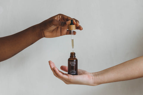 Hand holding dropper and letting a single drop fall into a tincture bottle held by another hand