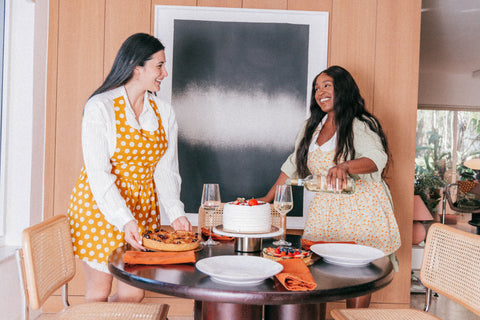 Two women in retro aprons setting a fruit tart on a kitchen table and pouring white wine into wine glass