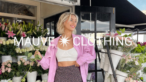 Smiling, blond woman wearing a pink button up shirt and holding a single rose with the Mauve Clothing logo in foreground